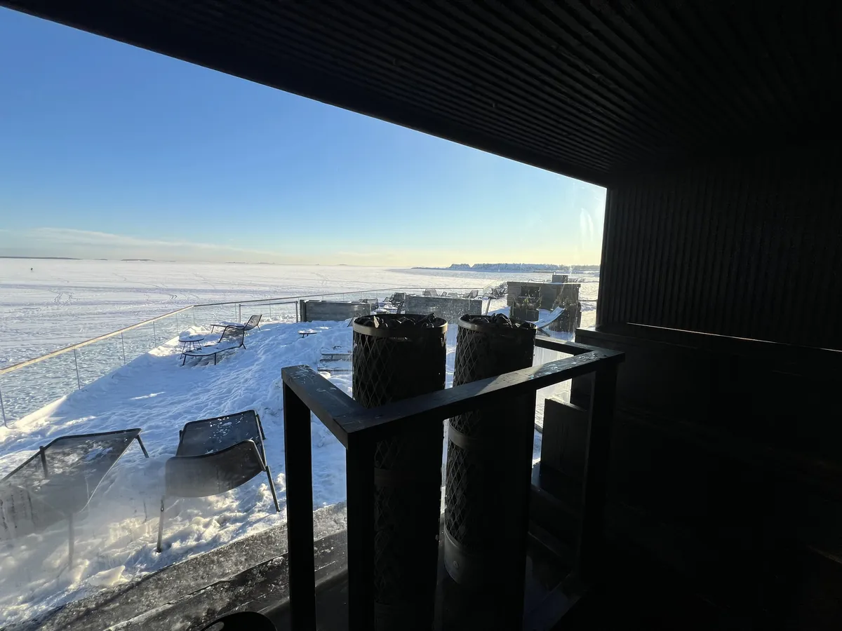 暗いサウナ施設の室内から、凍った海と雪のテラスを見る。手前に金網に入ったサウナストーンの柱が二本立つ