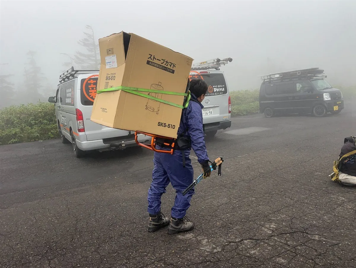 霧の山頂駐車場で、新しいストーブの箱を背負子で背負って出発するスタッフ