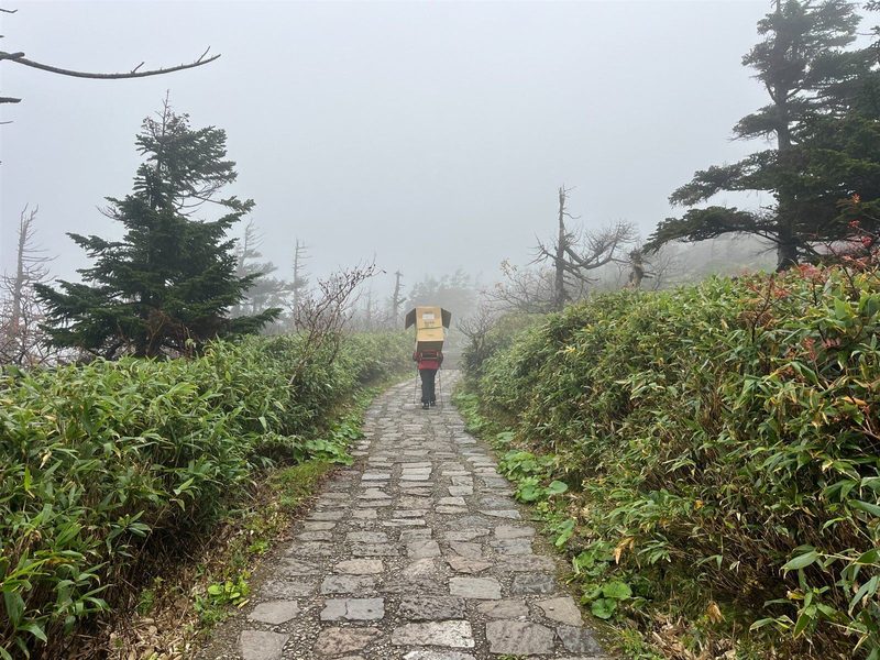 霧に包まれた八幡平の石畳の登山道を、ストーブを背負って運ぶ