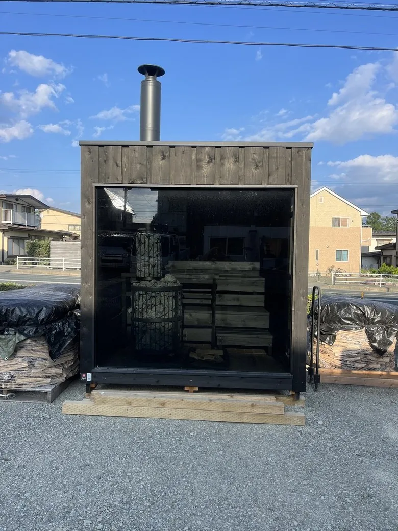 青空の下、黒い板張りのサウナ小屋。ガラス面ごしに石を積んだヒーターとベンチが見える(当社ショールーム)