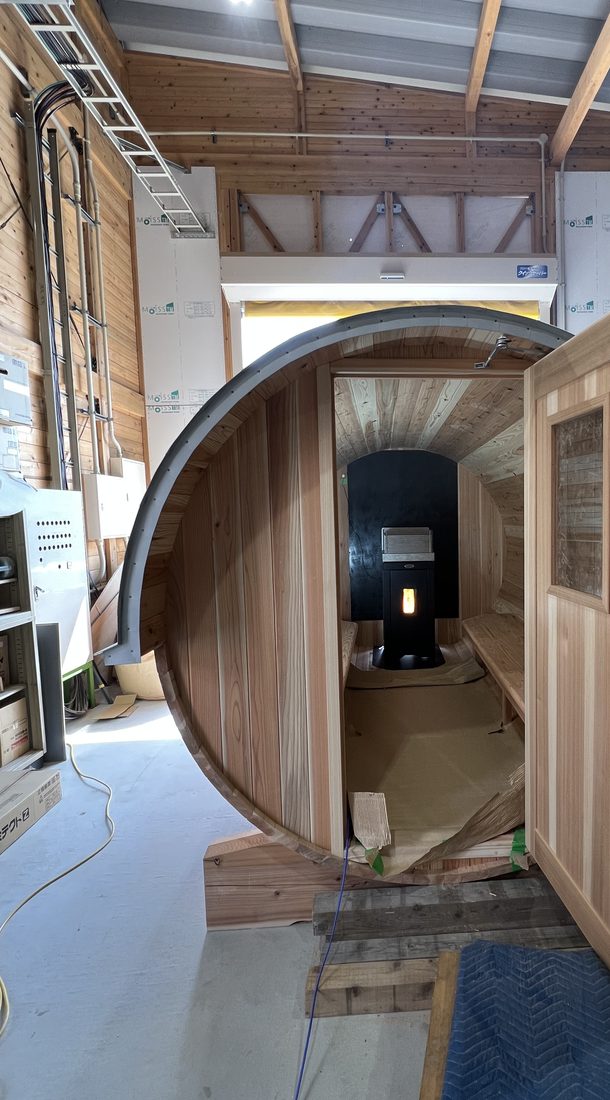 当社施工：作業場で扉を開けたバレルサウナ。奥に黒いペレットサウナヒーターが見える