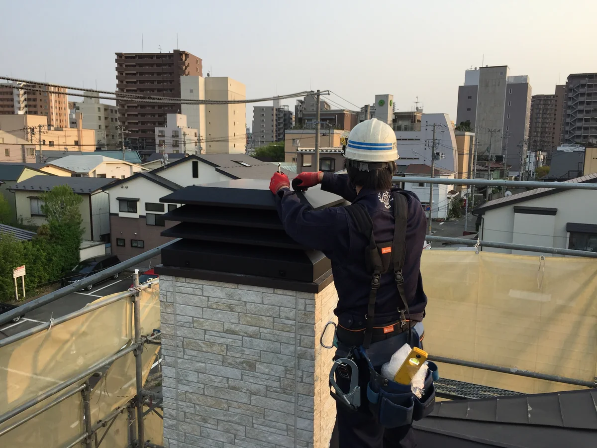 屋根の上で、石積み調の煙突の天端を押さえているところ。腰に工具袋と安全帯をつけている