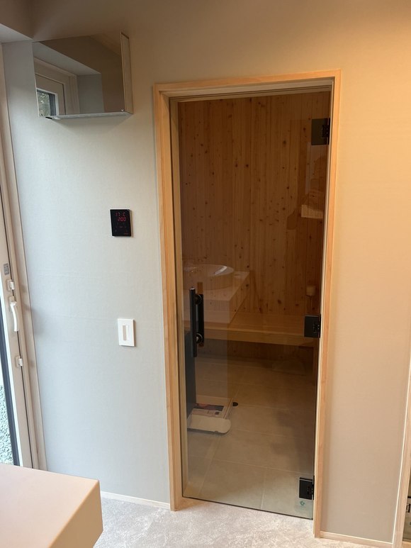 当社施工：脱衣室からサウナ室のガラス扉を見たところ。壁に黒いコントローラーと照明スイッチ